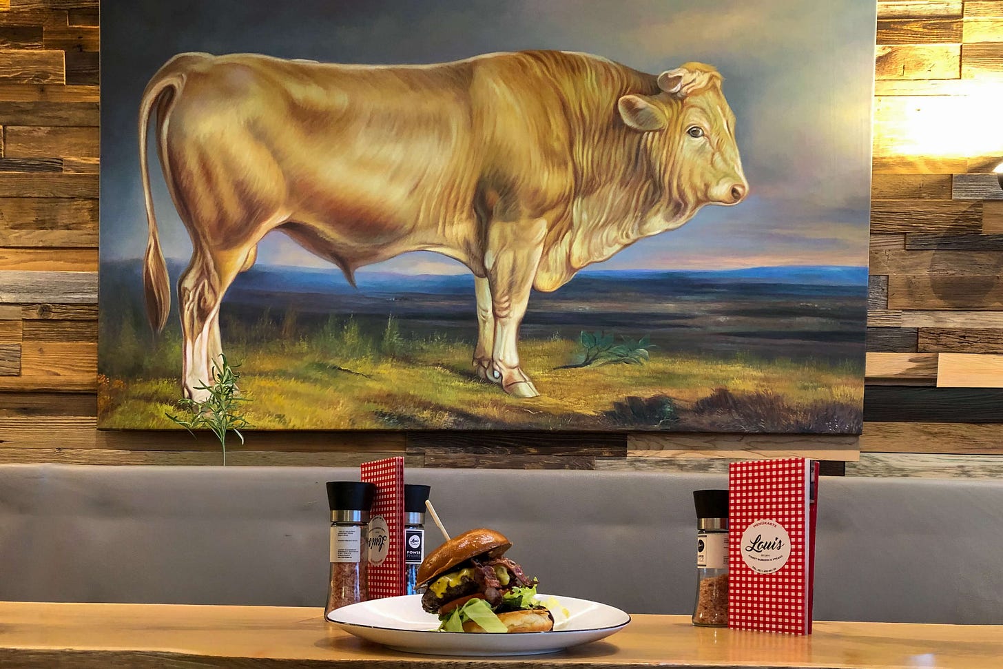 Innenraum eines Restaurants mit rustikaler Holzwand, an der ein grosses Gemälde eines hellbraunen Stiers in Landschaft hängt. Davor steht auf einem Tisch ein reich belegter Burger auf einem weissen Teller, flankiert von zwei Gewürzmühlen und rot-weiss karierten Speisekarten. Innenraum eines Restaurants mit rustikaler Holzwand, an der ein grosses Gemälde eines hellbraunen Stiers in Landschaft hängt. Davor steht auf einem Tisch ein reich belegter Burger auf einem weissen Teller, flankiert von zwei Gewürzmühlen und rot-weiss karierten Speisekarten.