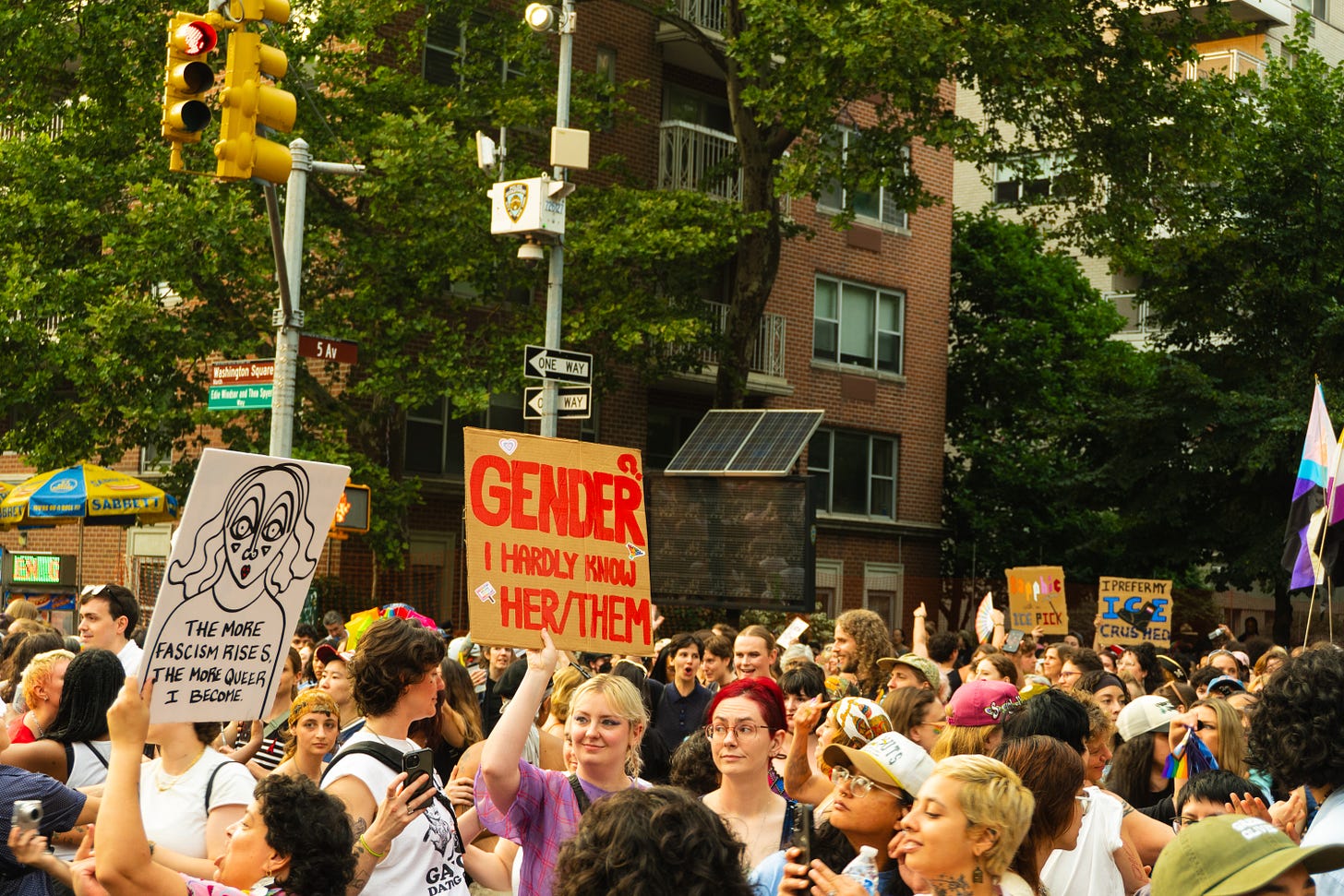 A crowd of queers under a green understory of trees. In the center, someone in a purple shirt and blonde hair lightly smiles and holds up a cardboard sign reading “Gender? I hardly Know Her/Them”