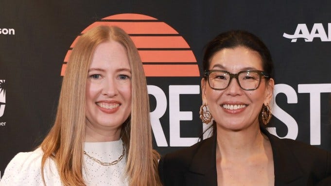 Two femmes, one white with blond hair and one Taiwanese with dark hair, are smiling on a step and repeat line at what seems like a movie premiere.
