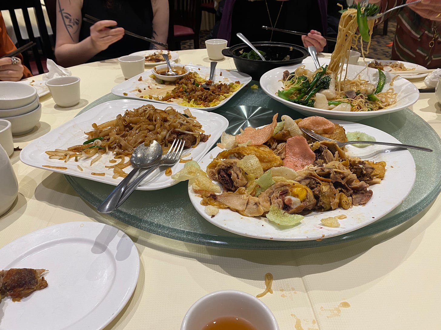 Several large plates of partially-eaten noodles and meat on a glass lazy susan, forks and serving spoons resting on them. People's hands and arms are visible in the background.