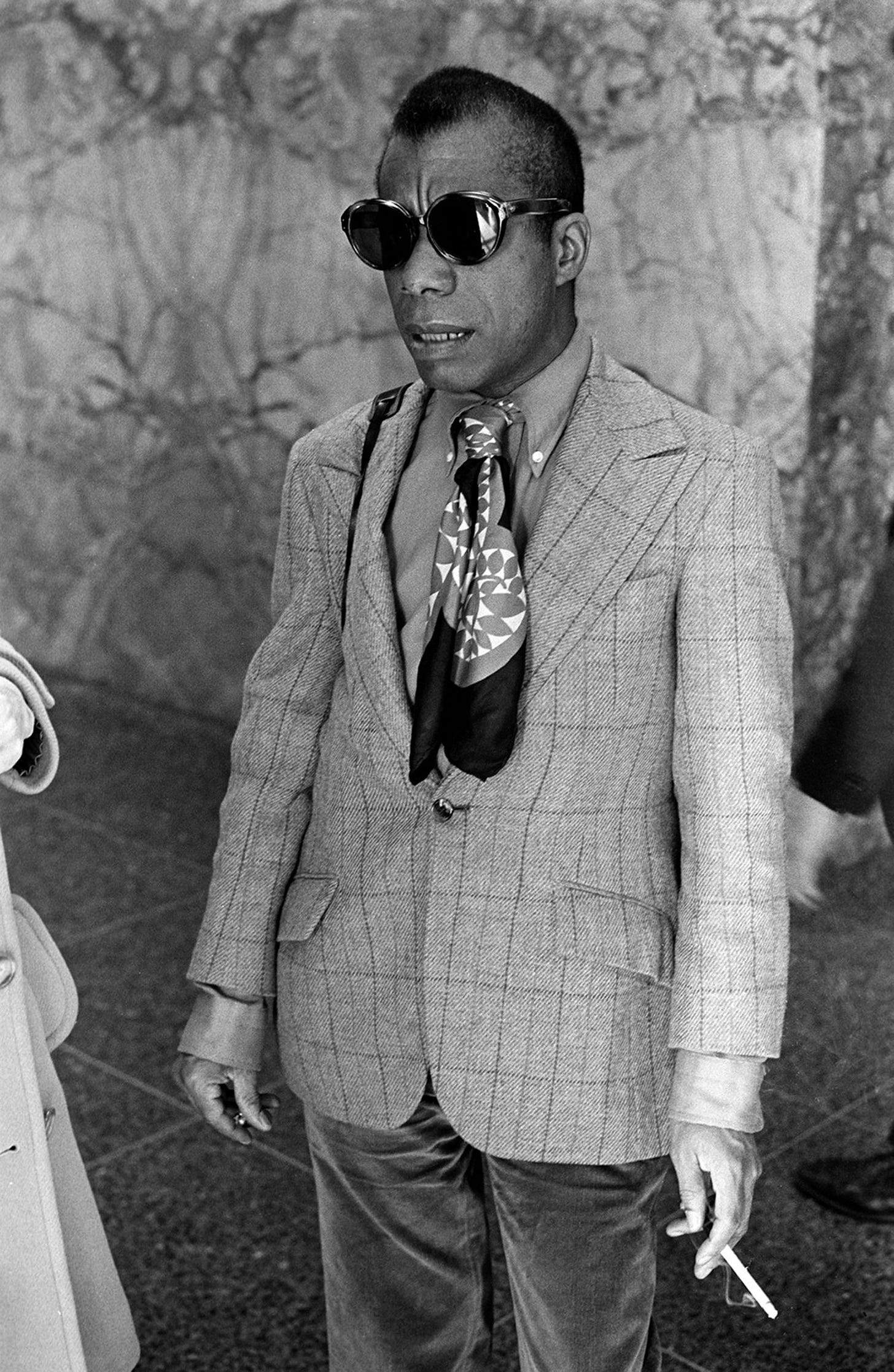 Black-and-white photograph of James Baldwin standing confidently in a checkered blazer, wide-lapel shirt, and bold patterned scarf. He wears oversized sunglasses and holds a cigarette in one hand. His expression is composed and observant, exuding intellect and cool defiance. The textured marble wall in the background adds a formal, urban backdrop to his effortlessly stylish presence.