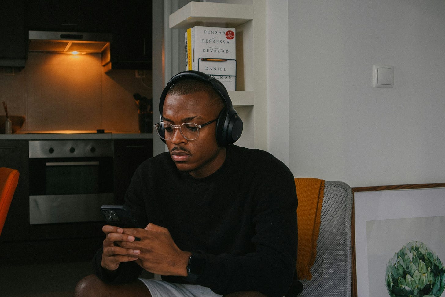 Young man wearing headphones and glasses, seated indoors, looking at a smartphone. A kitchen and books are visible in the background. Photo by Nubelson Fernandes on Unsplash