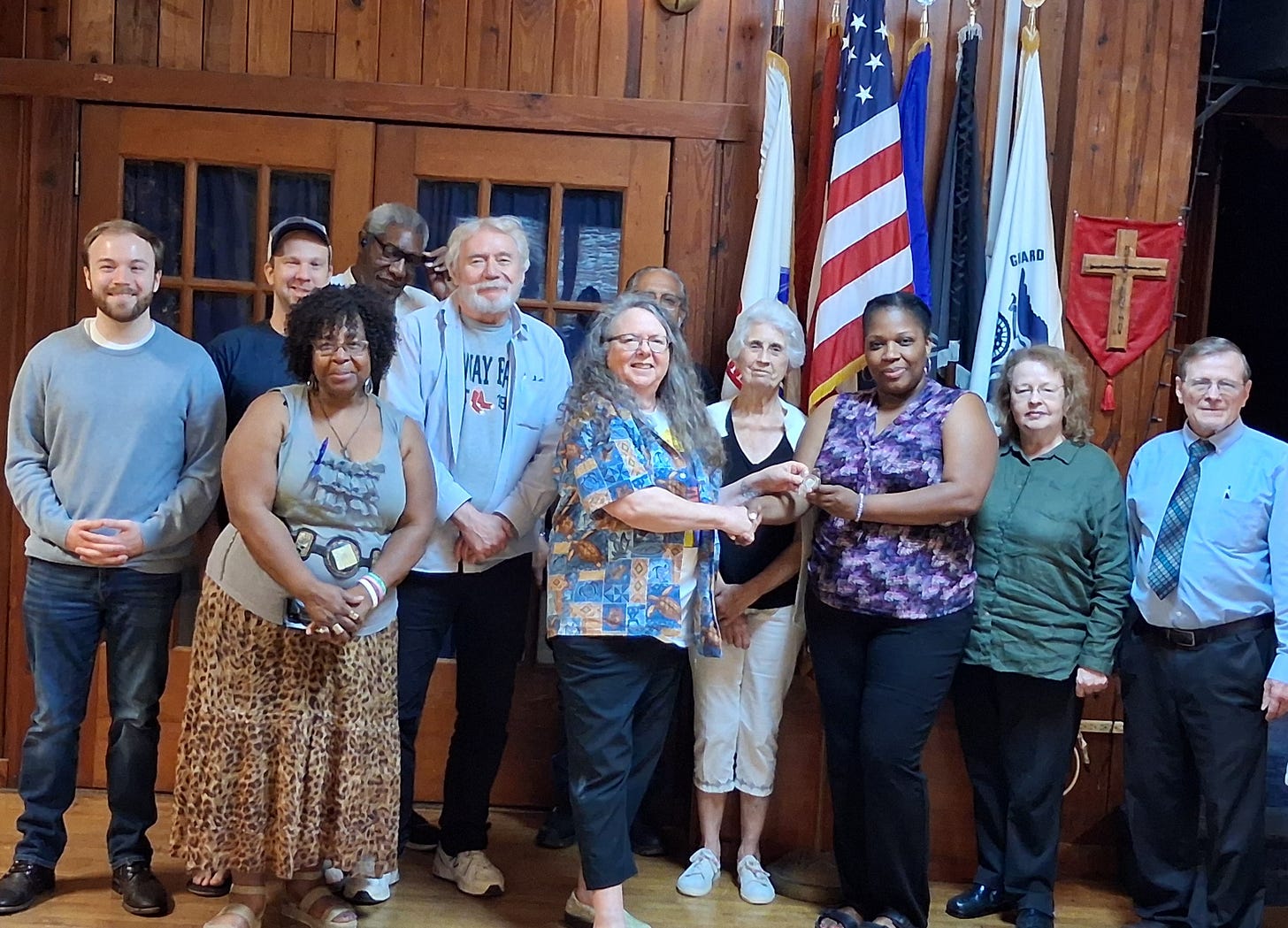 A group of adults poses for a photo inside a wood-paneled hall, with two women in front shaking hands while holding a small item. Behind them are several flags, including the United States flag, and a red banner with a wooden cross. One man wears a shirt with “Fenway Park” and a Boston Red Sox logo.