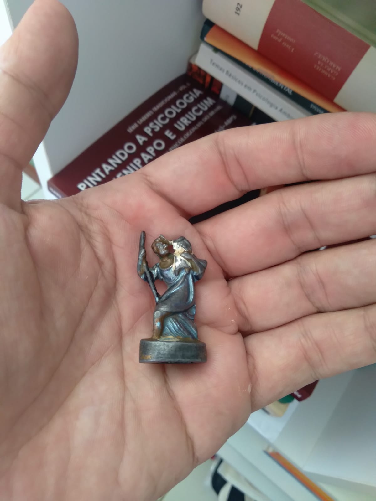 Close em minha mão espalmada. No entro dela, uma imagenzinha de chumbo de São Cristóvão, já desgastada pelo tempo, o menino Jesus a seu ombro sem a cabeça; ao fundo, pilhas de livros em uma estante, dos quais só se pode ler um pedaço do título de um: "Pintando a psicologia de jenipapo e urucum"