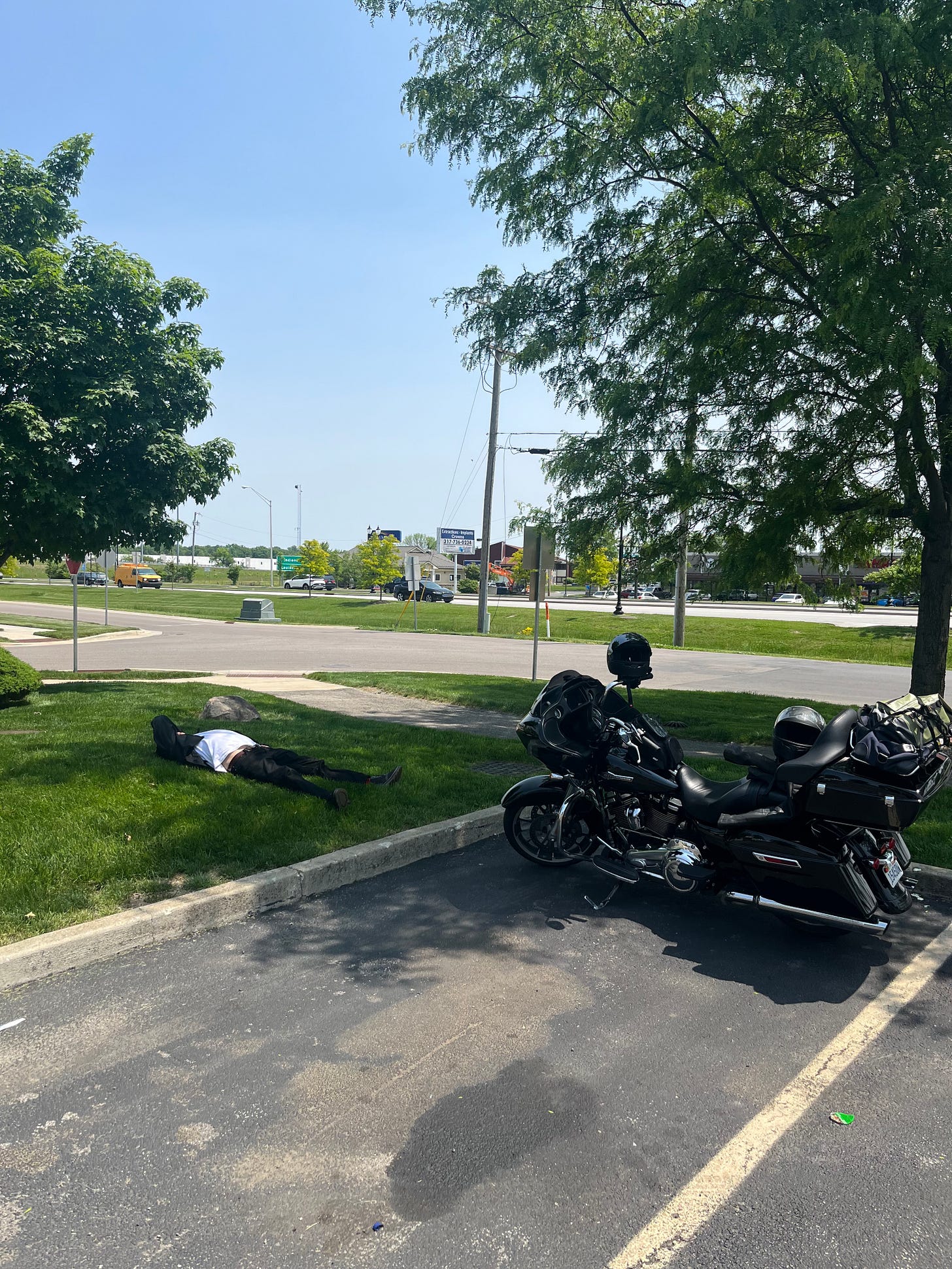 The Harley is parked in a parking spot. On a small patch of grass nearby, Derrick is lying faceup with his arm over his head.