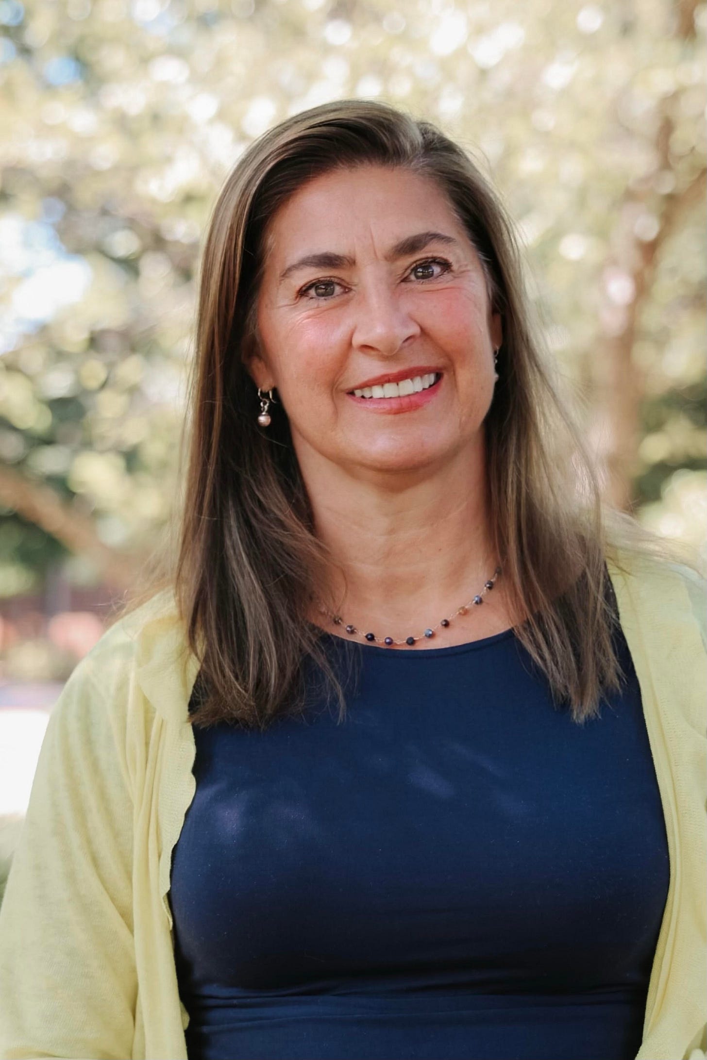 Leslie Manookian smiles outdoors in a navy top and yellow cardigan, trees blurred behind her. Leslie Manookian smiles outdoors in a navy top and yellow cardigan, trees blurred behind her.
