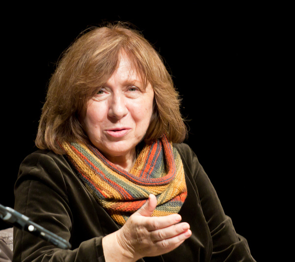 Foto de  Elke Wetzig/Wikipedia. Descrição de Imagem: a foto retrata Svetlana Aleksiévitch. O fundo é preto. Ela é uma senhora branca com cabelos loiros, quase ruivos, na altura dos ombros. Ela está falando e uma das mãos está gesticulando. Ela usa uma blusa de manga comprida de tom escuro e um lenço ao redor do pescoço, ele parece ter sido feito manualmente e é colorido, cheio de cores vivas, como laranja, verde, roxo, azul e verde.  