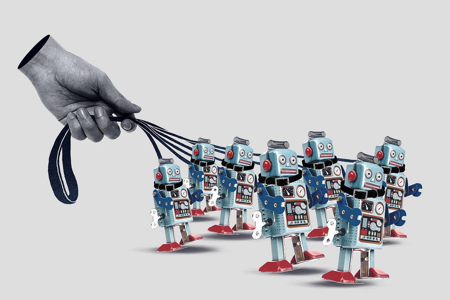A large grayscale human hand holds multiple leashes, each attached to a retro-style toy robot. The robots are metallic blue with red feet, wind-up keys, and analog dials on their chests, arranged in a group as if being walked like pets. The background is a plain light gray, emphasizing the surreal and symbolic nature of the scene, which conveys themes of control, automation, or conformity in technology.
