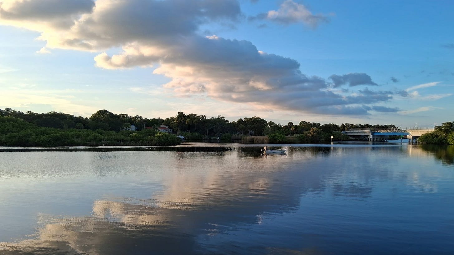 A wide, peaceful view of Turkey Creek in Palm Bay, Florida, showing a small boat on glassy, reflective water with a bridge visible in the distance under scattered afternoon clouds.