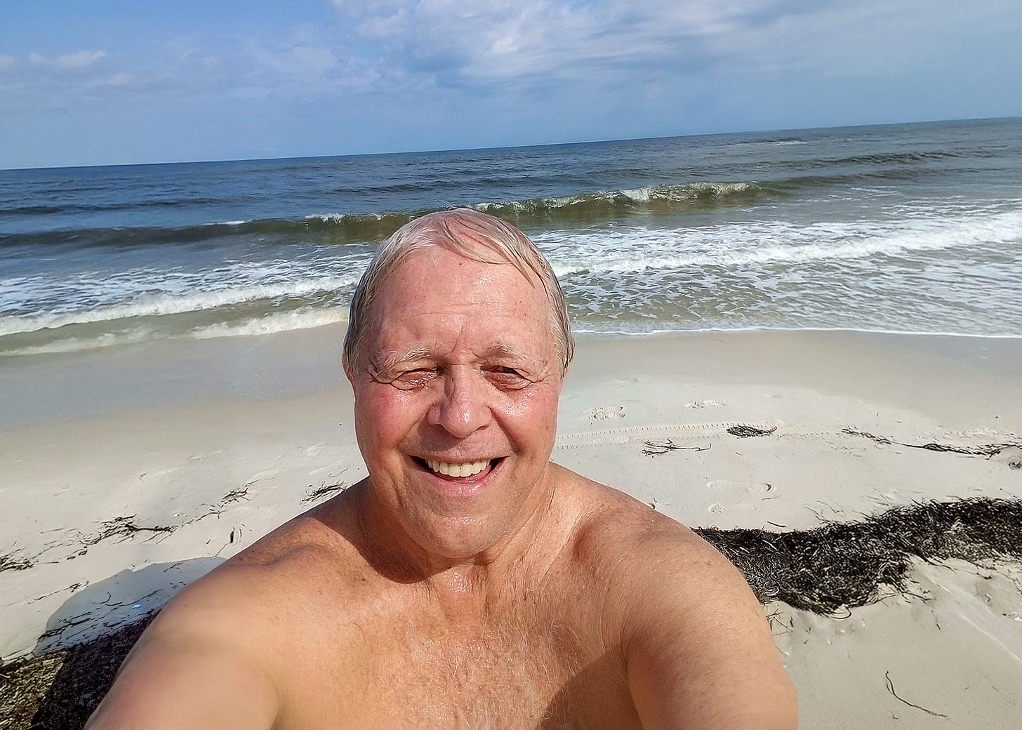 Bruce Frendahl smiling in a nude beach selfie, standing on a sandy shoreline with ocean waves behind him under a partly cloudy sky.