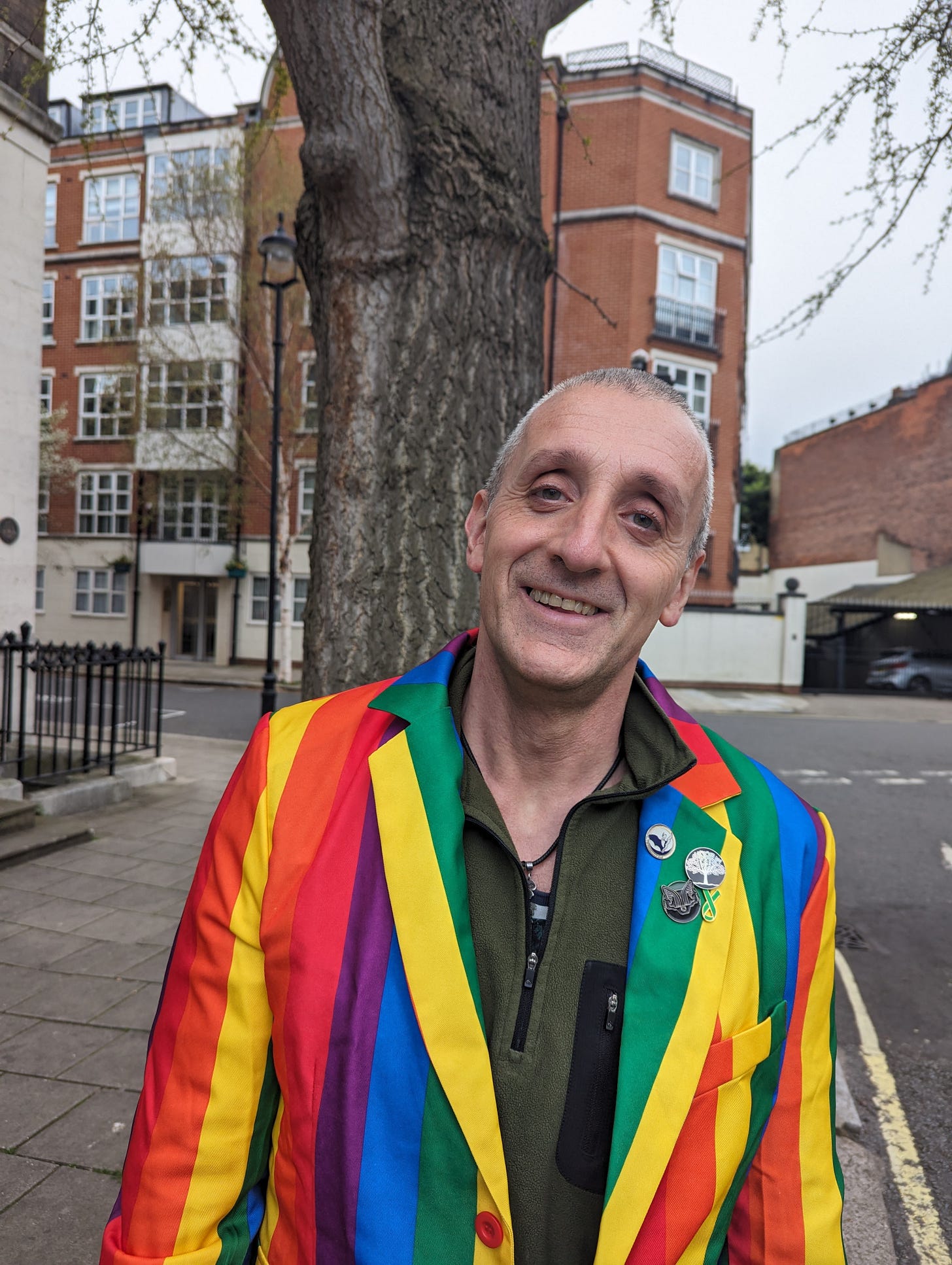 A man with short greying hair and kind green-ish eyes smiles at the camera, wearing a rainbow jacket A man with short greying hair and kind green-ish eyes smiles at the camera, wearing a rainbow jacket