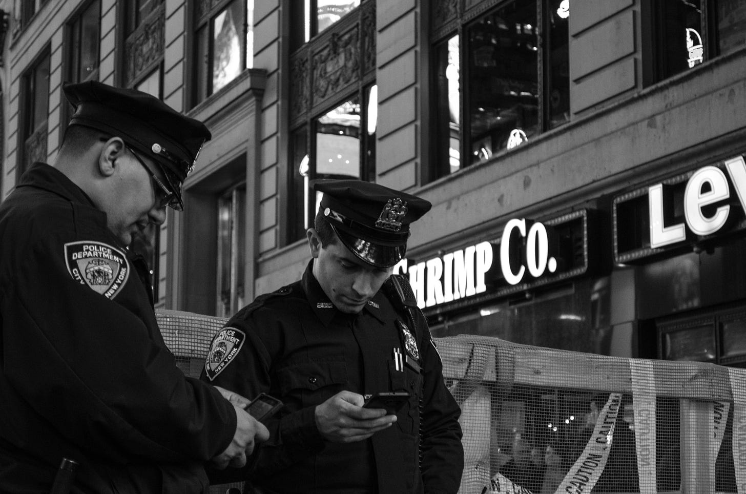 A black and white photo of two uniformed NYPD police officers looking down at their phones on a public street.