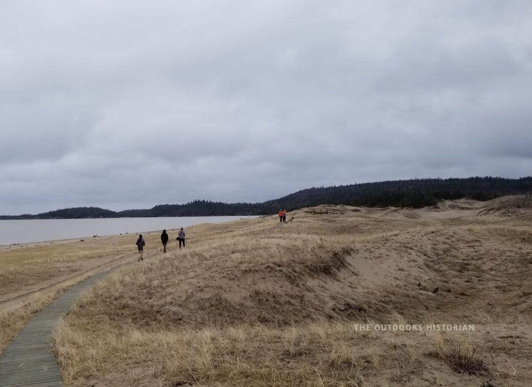 A wide, low-angle shot of a wooden boardwalk winding through gradual, grass-covered sand dunes on a stormy May day. A small group of students, seen from behind, walk along the path in Biigtigong Nishnaabeg territory. The sky is a heavy, unsettled grey, and the dunes are low and subtle compared to the massive cliffs of the South Shore, showing the sparse vegetation of a rare boreal dune ecosystem.