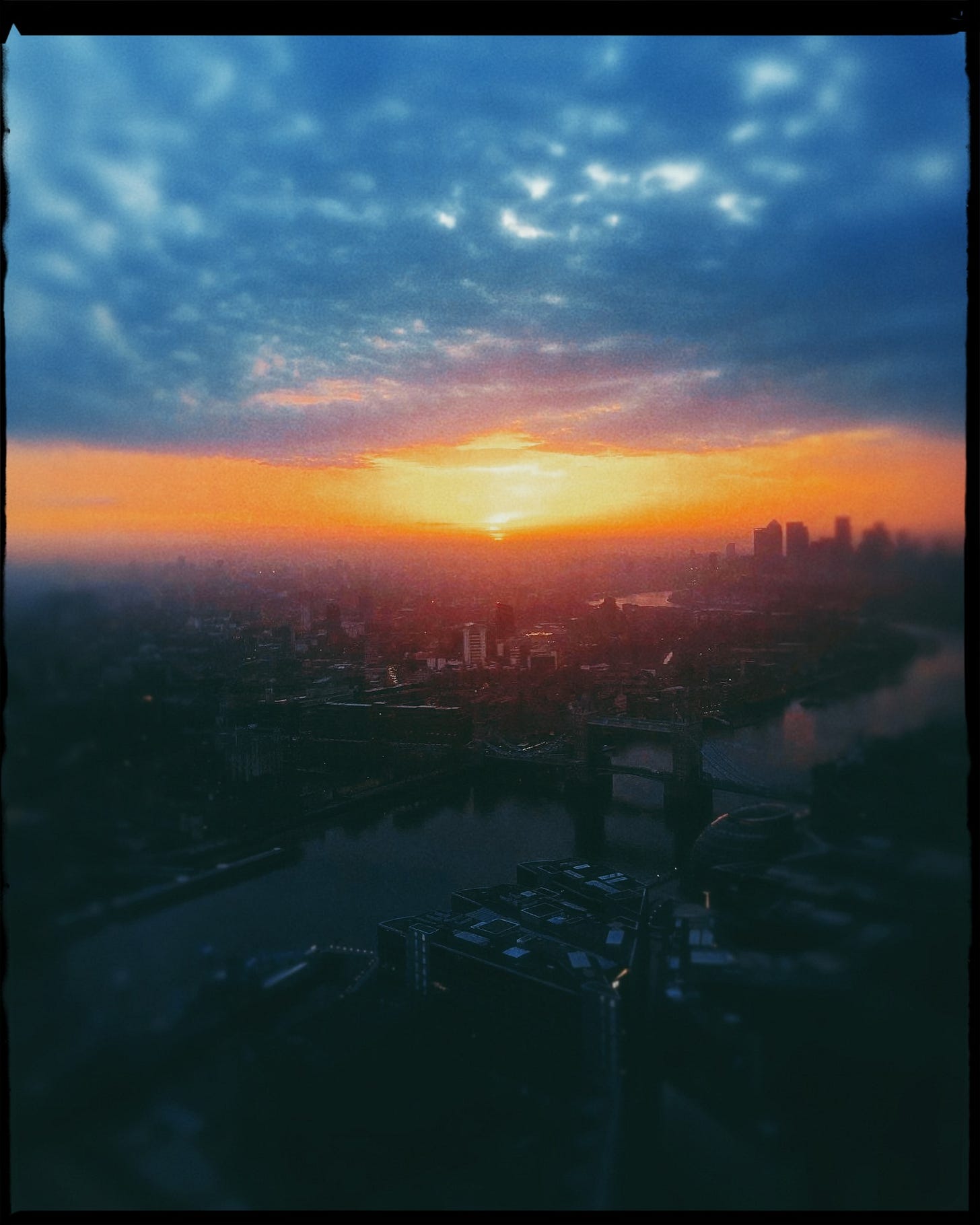 A view of London at sunrise, taken from the Shard, overlooking Tower Bridge and the Thames. There is a hazy filter on the photo so the details are hard to make out, but the colour palette is bold: blue fluffy clouds, an orange stripe of sky, and a bright yellow sun just starting to peek over the horizon.