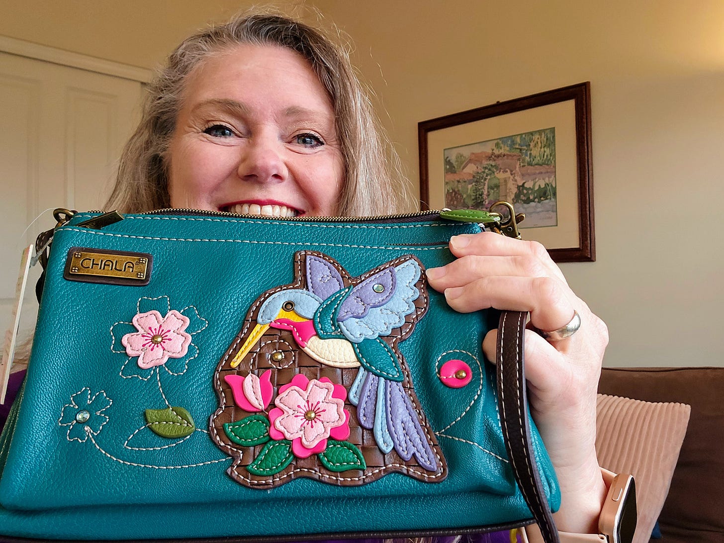 Theresa Carter Goodrich holding a purse stitched with a hummingbird. In the background is a pastel painting.