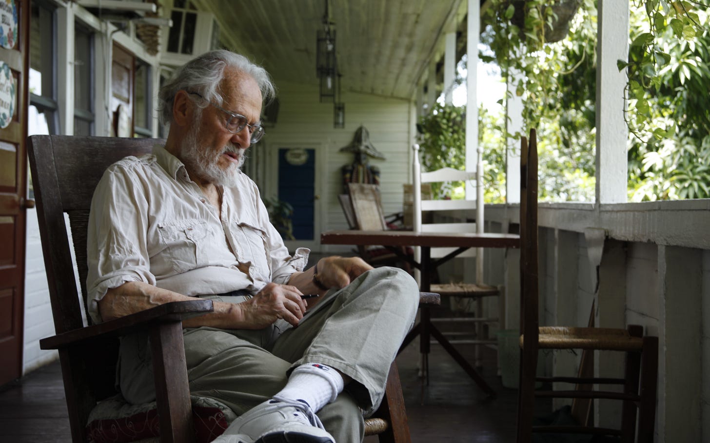 Herbert Gold, white-haired and bearded, sits reading in a wooden rocking chair on the shaded veranda of the Hotel Oloffson in Port-au-Prince, with white gingerbread woodwork and green foliage beyond. Herbert Gold, white-haired and bearded, sits reading in a wooden rocking chair on the shaded veranda of the Hotel Oloffson in Port-au-Prince, with white gingerbread woodwork and green foliage beyond.
