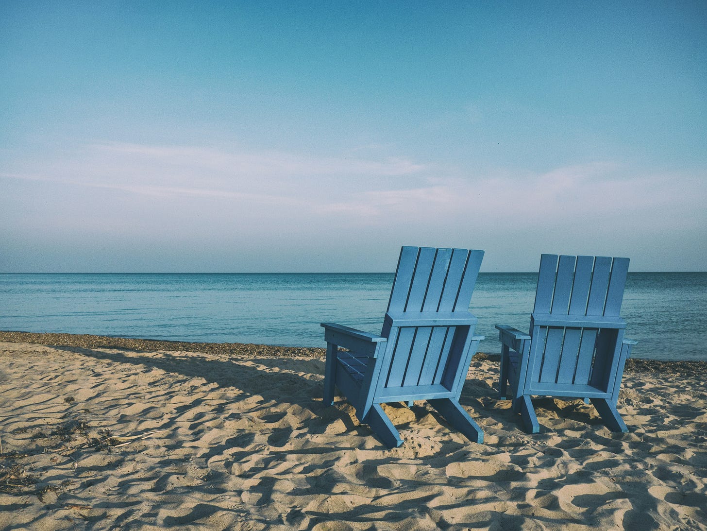 two blue wooden chairs sitting on the beach near the blue water under a blue sky