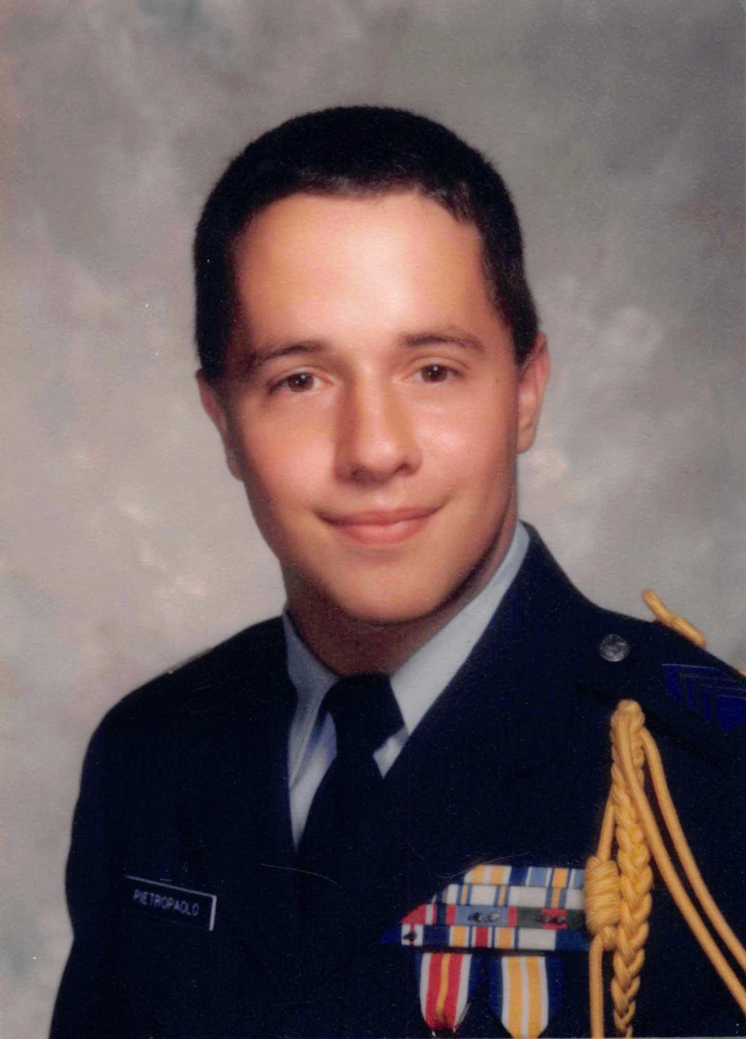 Tony in a dark blue Air Force Junior ROTC dress uniform, photographed in a studio against a gray backdrop. He has short dark hair and a slight smile. His uniform has a light blue shirt and dark tie, a gold braided cord over the right shoulder, a row of colorful ribbons and medals on the chest, and a name tag reading PIETROPAOLO.