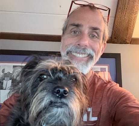 The image shows a person wearing a red t-shirt and holding a small, black-and-tan dog, with a wall and a framed picture in the background.
AI-generated content may be incorrect.