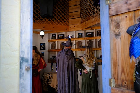 Bards singing in a tavern wearing medieval garb, an elf and wizard surveying goods at a shop, fire-twirler show, Geralt and another warrior deep in conversation, a man playing dead with an arrow under his armpit, guests walking toward the camera, a woman with a horse head looking over a table.