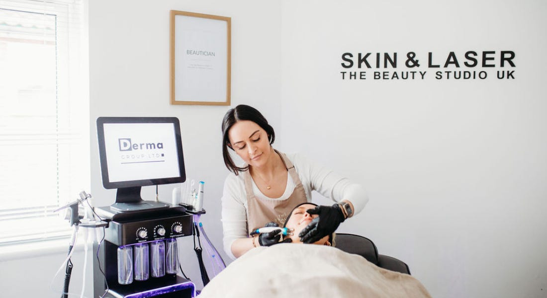 A woman giving a hydrafacial to someone lying down with a blanket over them, with a machine to the left and white walls behind A woman giving a hydrafacial to someone lying down with a blanket over them, with a machine to the left and white walls behind