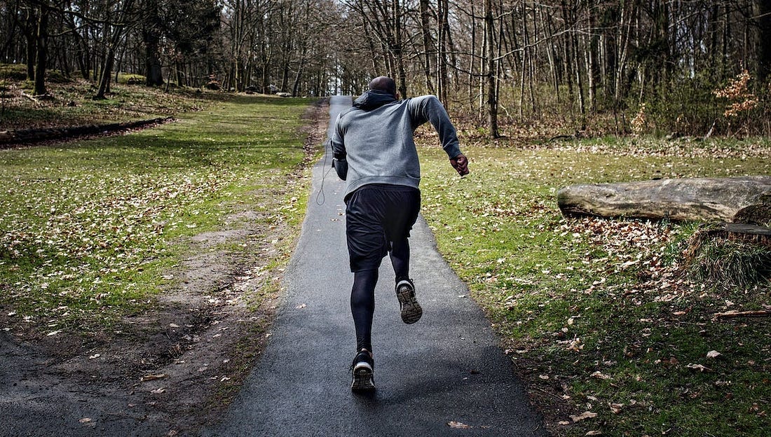 Full length rear view of determined male athlete jogging on narrow street in forest.