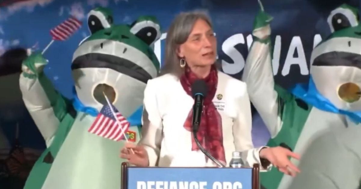A speaker addresses an audience at an event, flanked by inflatable frog figures holding American flags. A speaker addresses an audience at an event, flanked by inflatable frog figures holding American flags.