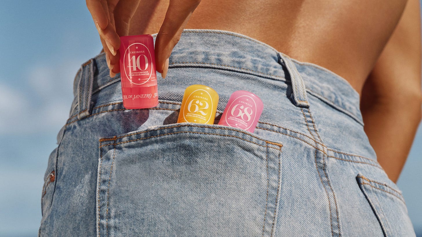 A close-up shot of a person wearing light-wash denim jeans, viewed from behind, standing under a bright blue sky. A hand is reaching out to pull a pink Sol de Janeiro '40' jelly perfume balm stick out of the back pocket. Two other balm sticks, '62' (yellow) and '68' (lighter pink), are also tucked into the same pocket.