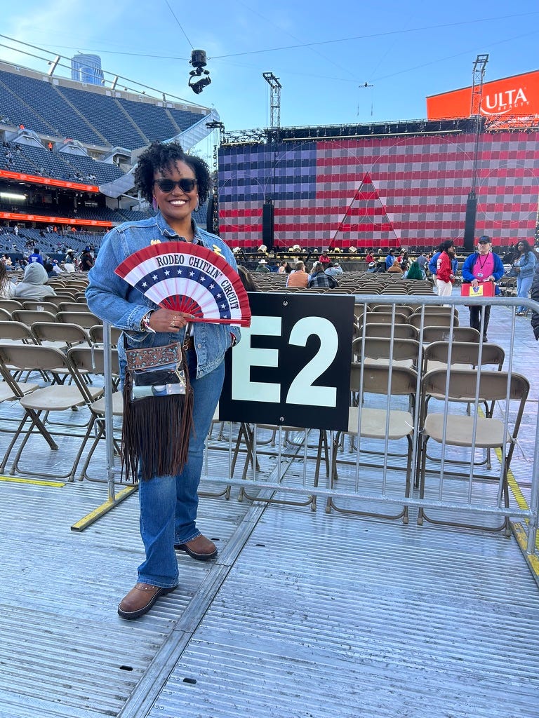 Me, a Black woman, standing in front of a screen showing the American flag, wearing a denim jacket and jeans, holding a red, white and blue fan that says Rodeo Chitlin Circuit