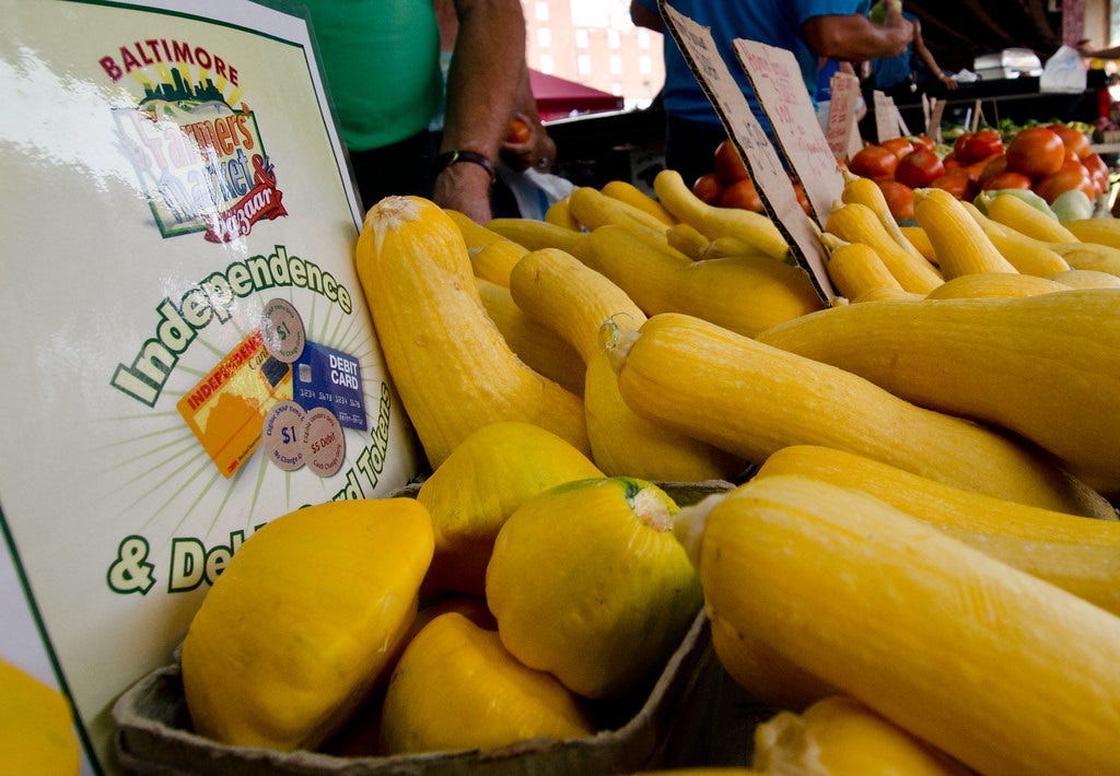 brightly colored vegetables (yellow squash in foreground, tomatoes and other produce behind) on a long table at a farmer's market. Next to the squash is a sign explaining that the vendor accepts debit cards and SNAP EBT payments