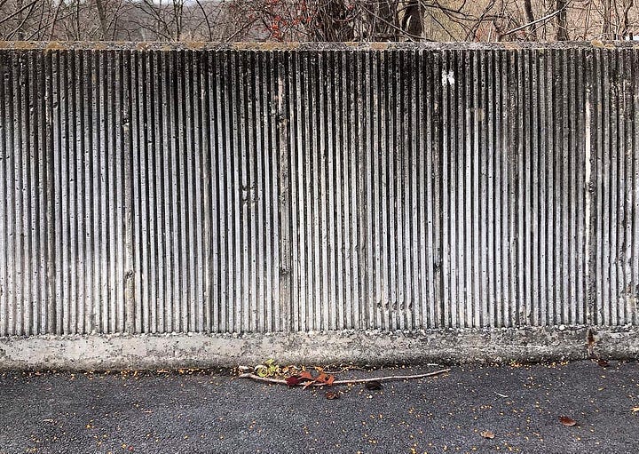 LEFT: Concrete wall next to the dentist office. The wall is made up of thin vertical grooves creating an interesting pattern as it weathered. Some of the vertical ridges have broken away in chunks. The top of the wall is stained dark with some moss growing on the top edge. RIGHT: Figure detail from NO SERVICE featuring a woman in yellow long-sleeved shirt with scoop neck and tight fitting jeans that end mid calf. She looks fashionable as she perches on a monolithic slab of concrete with her body turned as she holds out her camera.