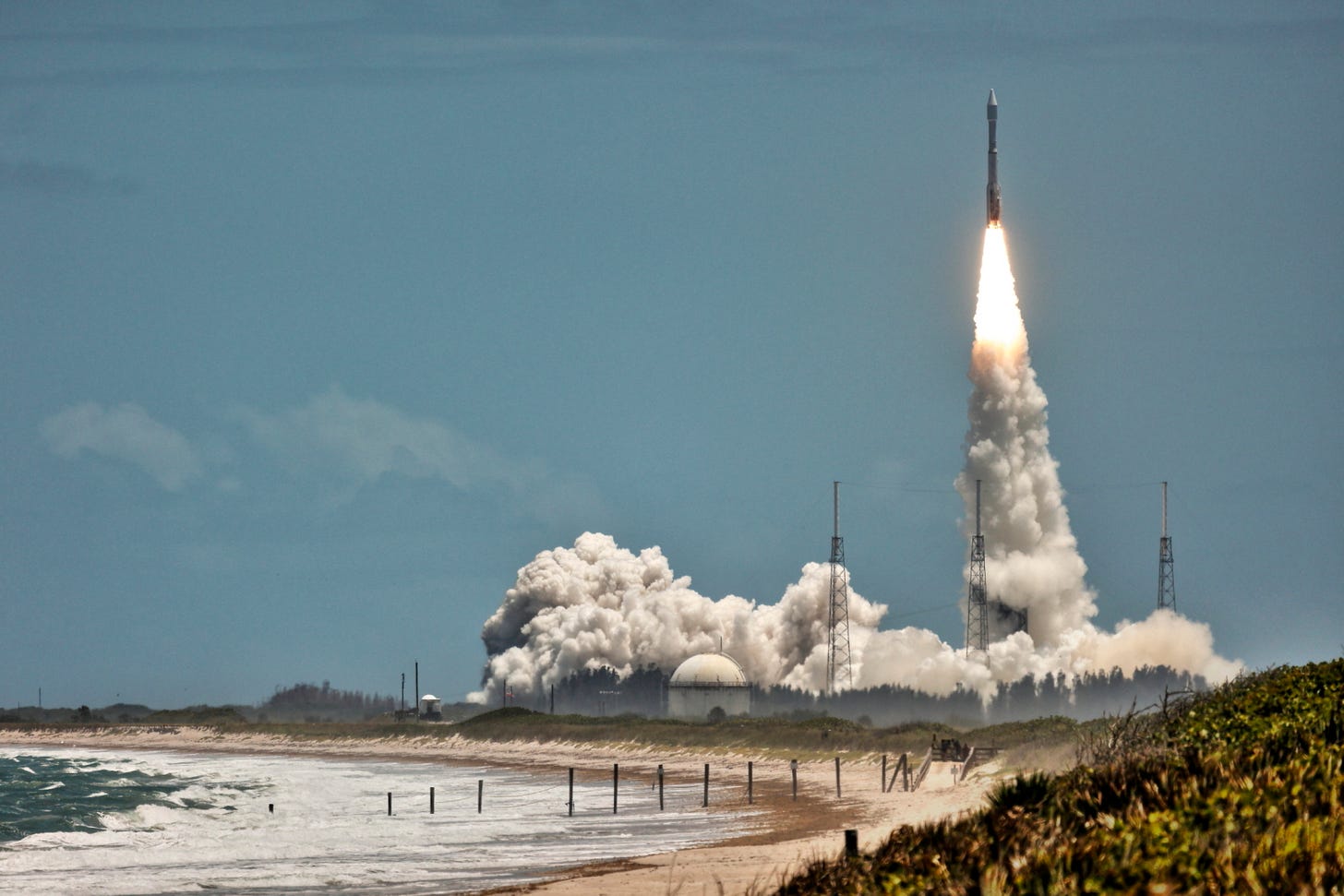 A rocket launches into a clear blue sky from Kennedy Space Center, with white smoke and flames trailing upward. In the foreground, waves crash along the shoreline of Playalinda Beach at Canaveral National Seashore. A rocket launches into a clear blue sky from Kennedy Space Center, with white smoke and flames trailing upward. In the foreground, waves crash along the shoreline of Playalinda Beach at Canaveral National Seashore.