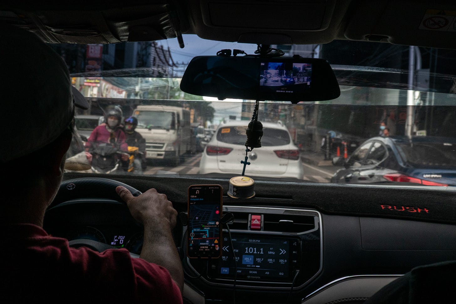 Photo from the back seat of a car, looking forward past the driver and through the windshield. The car is in Manila traffic - there are cars and scooter and trucks everywhere. The driver has both hands on the steering wheel. The road is narrow.