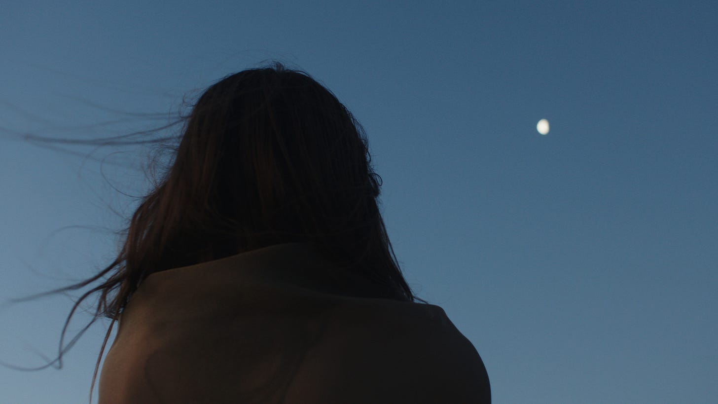 Seen from behind, a person with long hair lifted by the wind, their head and bare shoulders in shadow against a clear, deep blue dusk sky. Seen from behind, a person with long hair lifted by the wind, their head and bare shoulders in shadow against a clear, deep blue dusk sky.