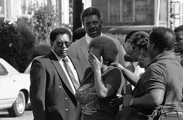 Mourners gather solemnly in front of the former MOVE headquarters, as the funeral procession for founder John Africa makes its way past. Mourners gather solemnly in front of the former MOVE headquarters, as the funeral procession for founder John Africa makes its way past.