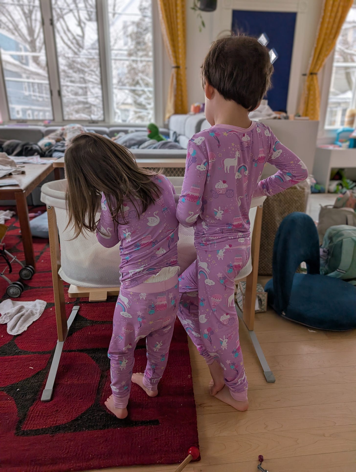 Photograph of two young children in purple pajamas in a cluttered house, looking down at a bassinet
