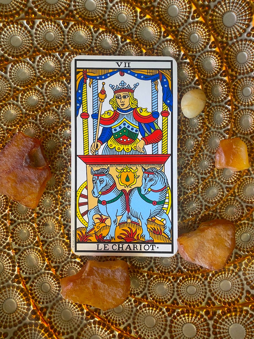 A Tarot card, number 7, the Chariot, sitting on a plate, with a swirling pattern around the outside, and unpolished rocks of carnelian around the card itself. The card shows a figure in a chariot, pulled by two horses.