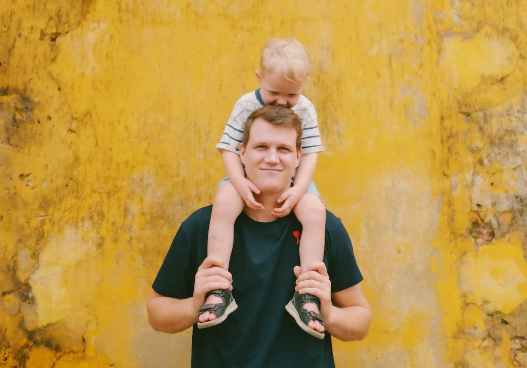Father carries son on shoulders against yellow wall.