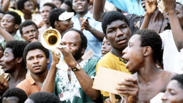 A crowd of spectators, a man in a traditional Nigerian shirt blows a trumpet, another sings, another looks directly at camera.