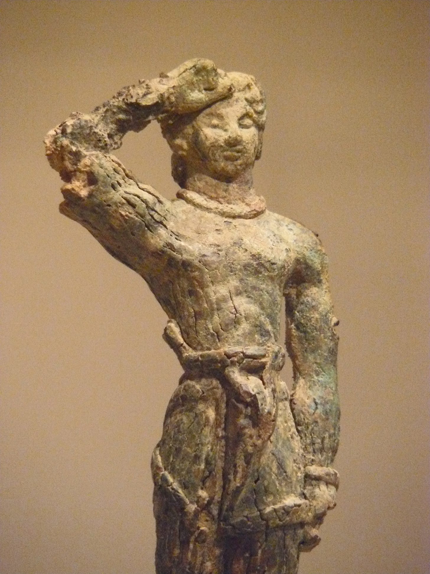 Bronze figurine of a young man with short hair, wearing a loincloth, posed in the Minoan salute, with his right arm raised so the back of his loosely-curled fist is against his forehead.