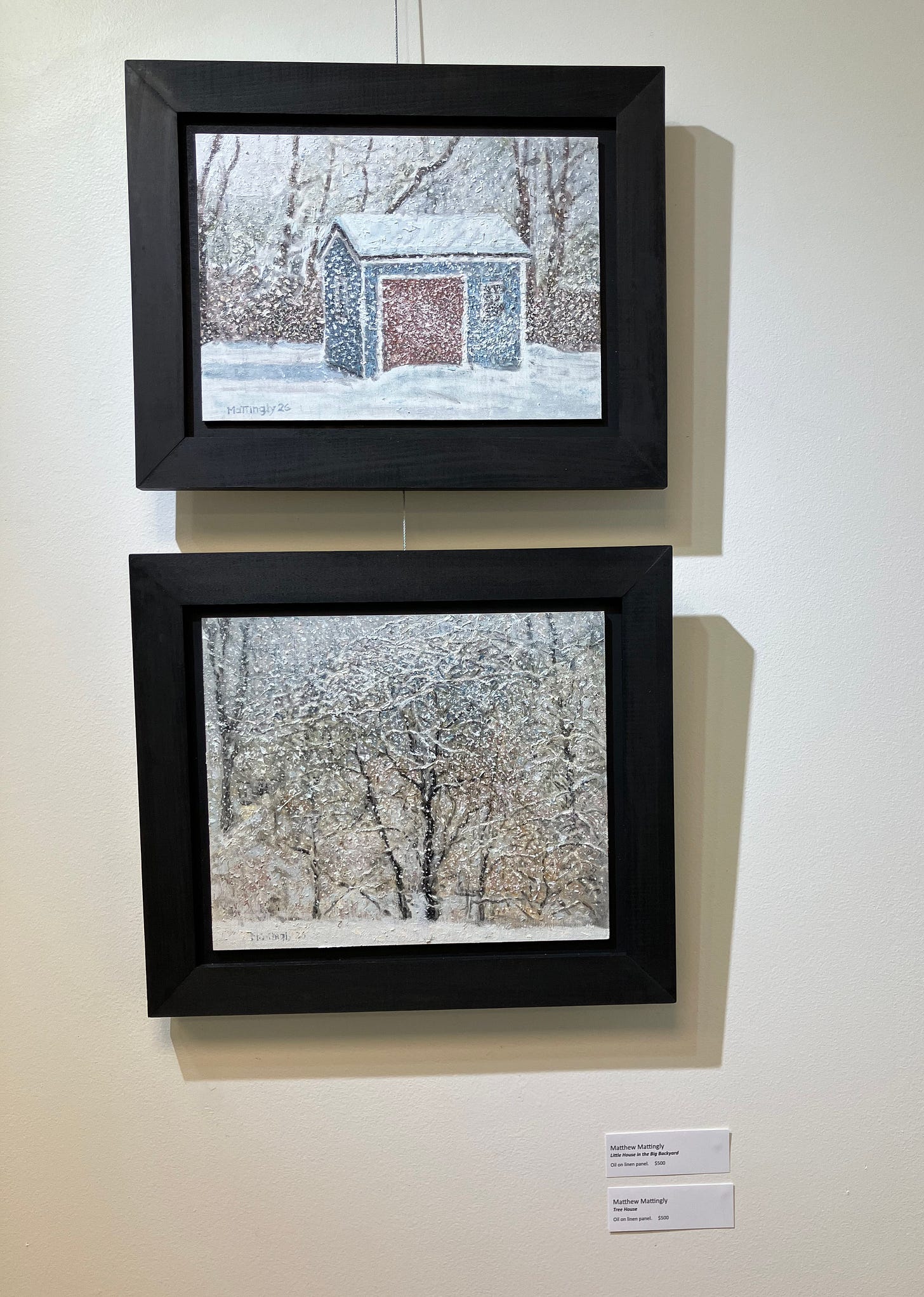 Two framed oil paintings hanging one above the other on white gallery wall. Top painting depicts a blue, red, and white garden shed surrounded by blowing snow against a backdrop of trees. The bottom painting shows a tree with intricate branches highlighted by snow amid a dense blizzard. Two framed oil paintings hanging one above the other on white gallery wall. Top painting depicts a blue, red, and white garden shed surrounded by blowing snow against a backdrop of trees. The bottom painting shows a tree with intricate branches highlighted by snow amid a dense blizzard.