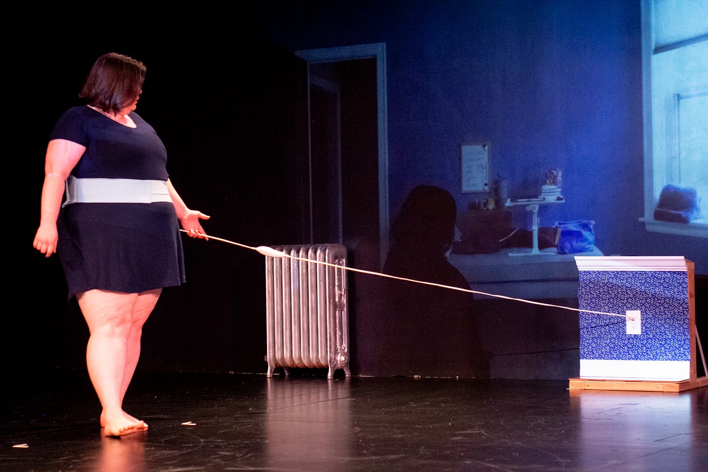 Maggie, a fat white woman in a short navy nightgown, walks in a semi-circle around a small wall. She’s connected to the wall via a heating pad wrapped around her stomach and plugged into a white outlet in the middle of the wall. In the background, a room with a grey chaise and turquoise walls is projected and a silver radiator sits against the projection.