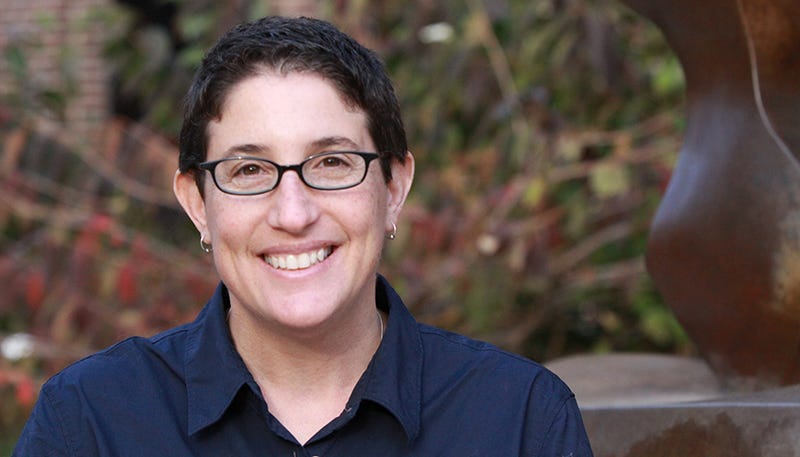 Laura's headshot, they are smiling looking directly at the camera, with black rimmed glases, short brown hair, white skin and a blue collared shirt.