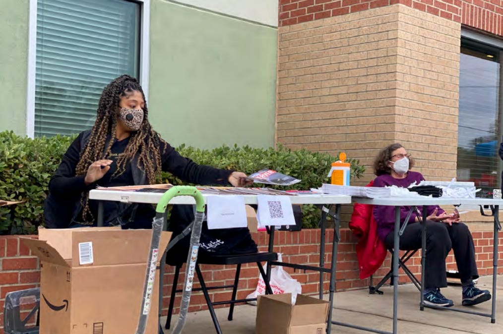 Zacharia Washington and a 21/7 member sit at the folding table in front of a green and brown building Zacharia Washington and a 21/7 member sit at the folding table in front of a green and brown building