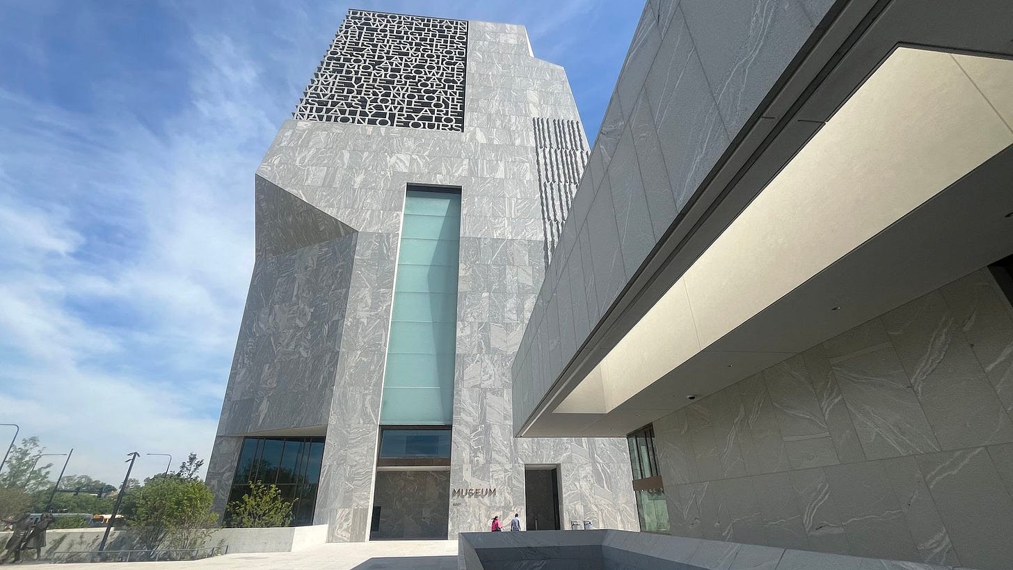 Angular grey marble museum with a black lattice panel of letters, a tall glass window, and a projecting overhang. Blue sky, entrance area with a few people and a visible MUSEUM sign.