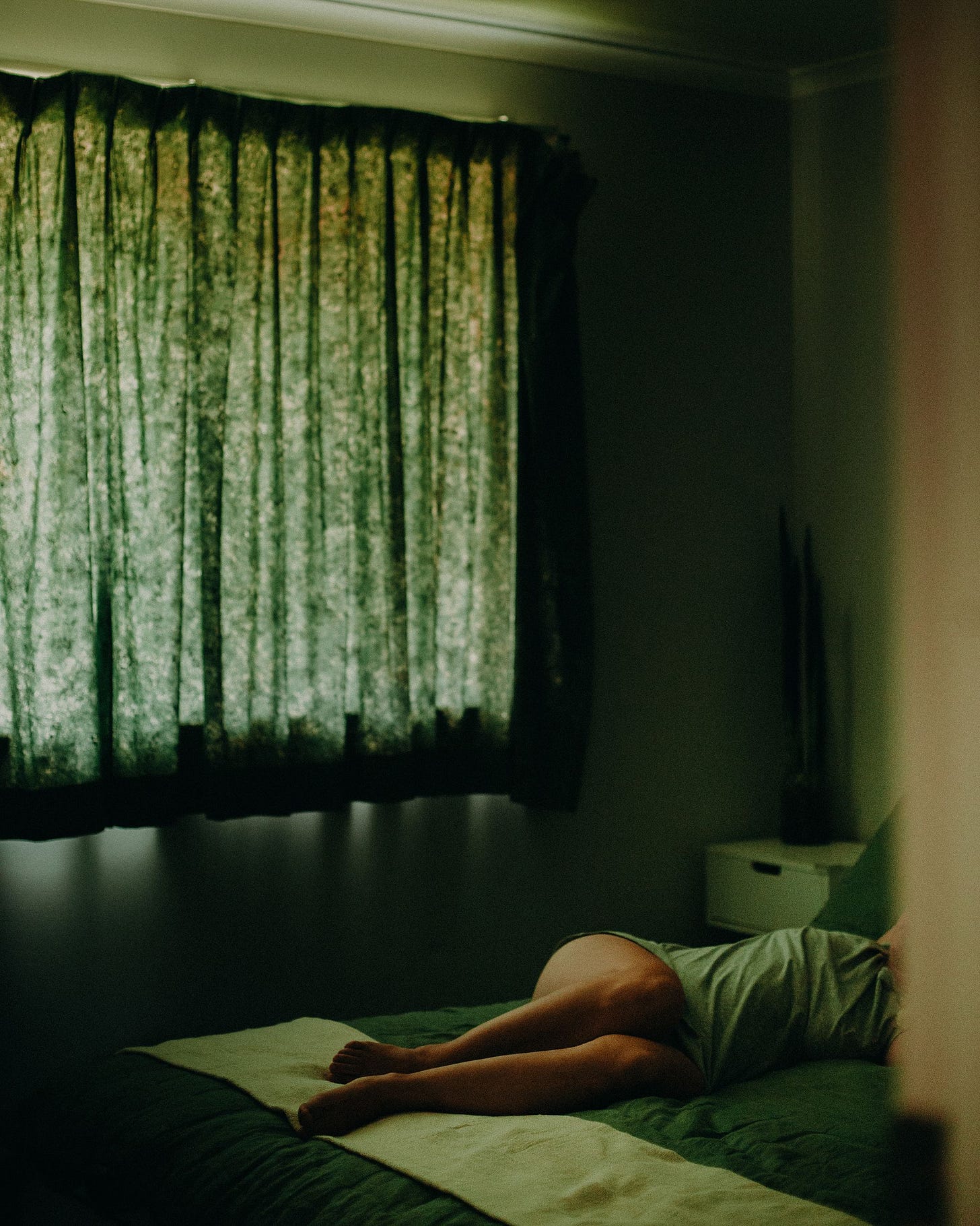 A person lies on a bed in a dimly lit room with green curtains and bedding A person lies on a bed in a dimly lit room with green curtains and bedding