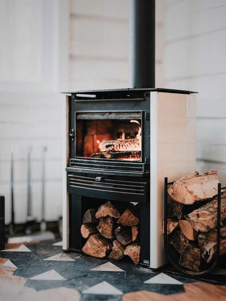 Soft domestic warmth during Imbolc: a wood-burning stove with stacked logs beside it, and a simple bowl of clear nourishing soup held mid-spoon, evoking heat, sustenance, and quiet restoration.