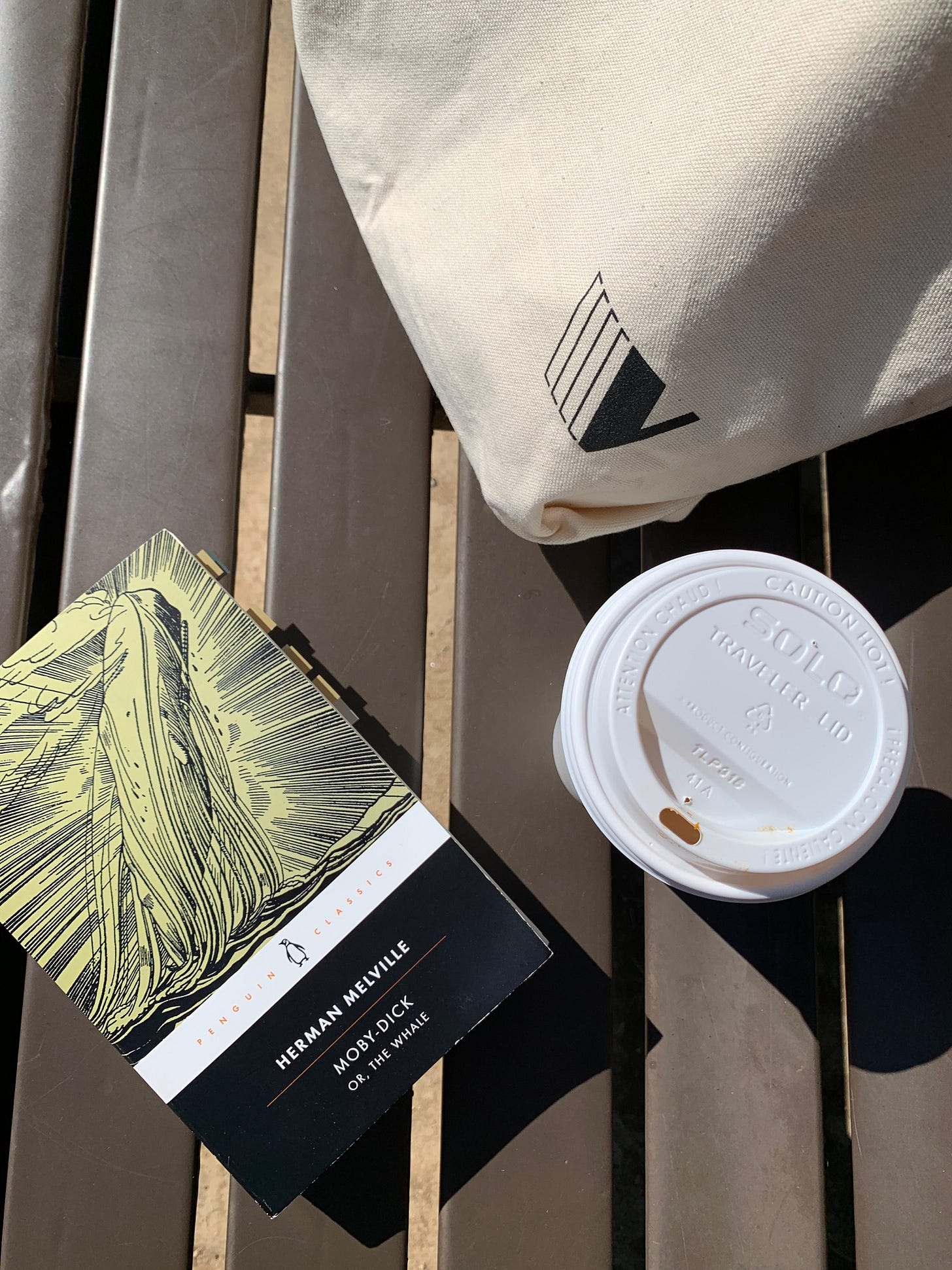 a copy of Moby Dick sits on a bench, next to a coffee cup and a tote bag.