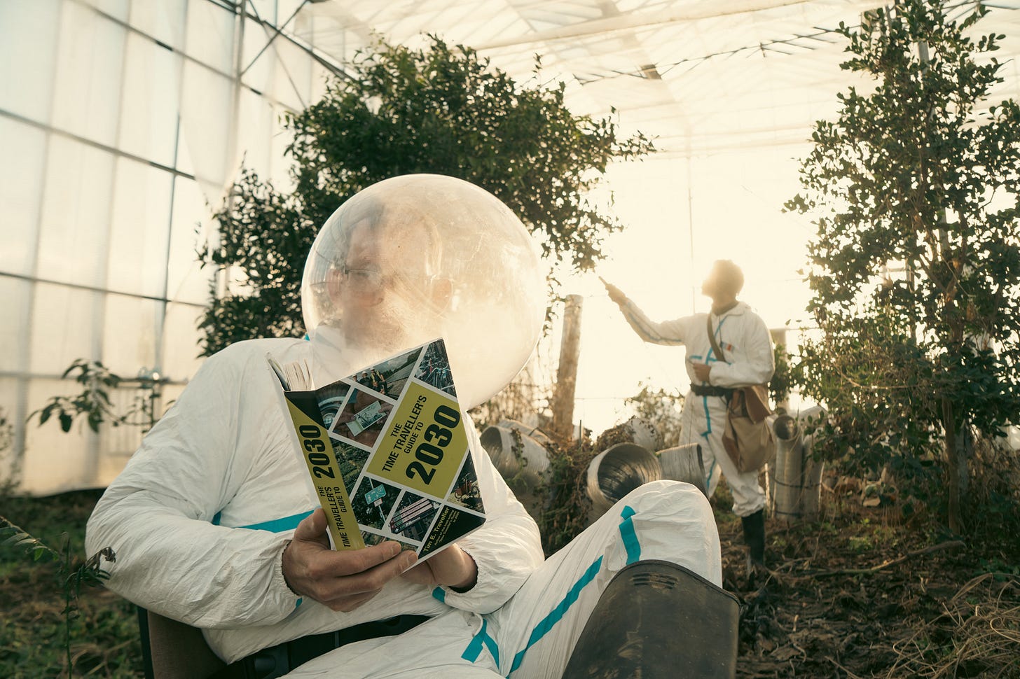 The author, Rob Hopkins, sits in a pretend space suit reading a book with the year 2030 prominently on the cover. In the background another person is recording sounds from a plant. They are both in a kind of enormous polytunnel. 