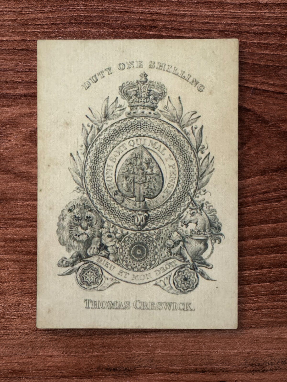 A close-up photograph of an antique "Old Frizzle" Ace of Spades playing card resting face up on a dark wooden surface. The highly ornate, engraved design features a crown at the top above the text "DUTY ONE SHILLING". In the center, a large spade symbol is enclosed by a garter reading "HONI SOIT QUI MAL Y PENSE". Below this, a lion and unicorn flank a banner that reads "DIEU ET MON DROIT". The name "THOMAS CRESWICK." is printed at the very bottom. A close-up photograph of an antique "Old Frizzle" Ace of Spades playing card resting face up on a dark wooden surface. The highly ornate, engraved design features a crown at the top above the text "DUTY ONE SHILLING". In the center, a large spade symbol is enclosed by a garter reading "HONI SOIT QUI MAL Y PENSE". Below this, a lion and unicorn flank a banner that reads "DIEU ET MON DROIT". The name "THOMAS CRESWICK." is printed at the very bottom.